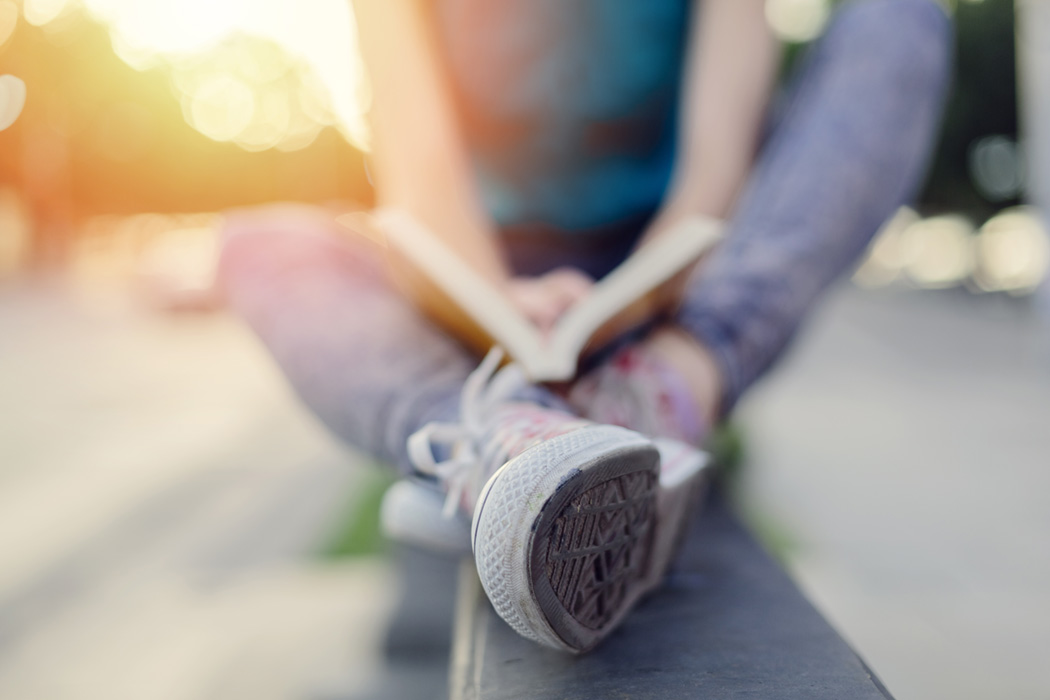 girl sitting along a wall with her sneakered feet facing camera, reading. Cropped to not include head, therefore age and features not important
