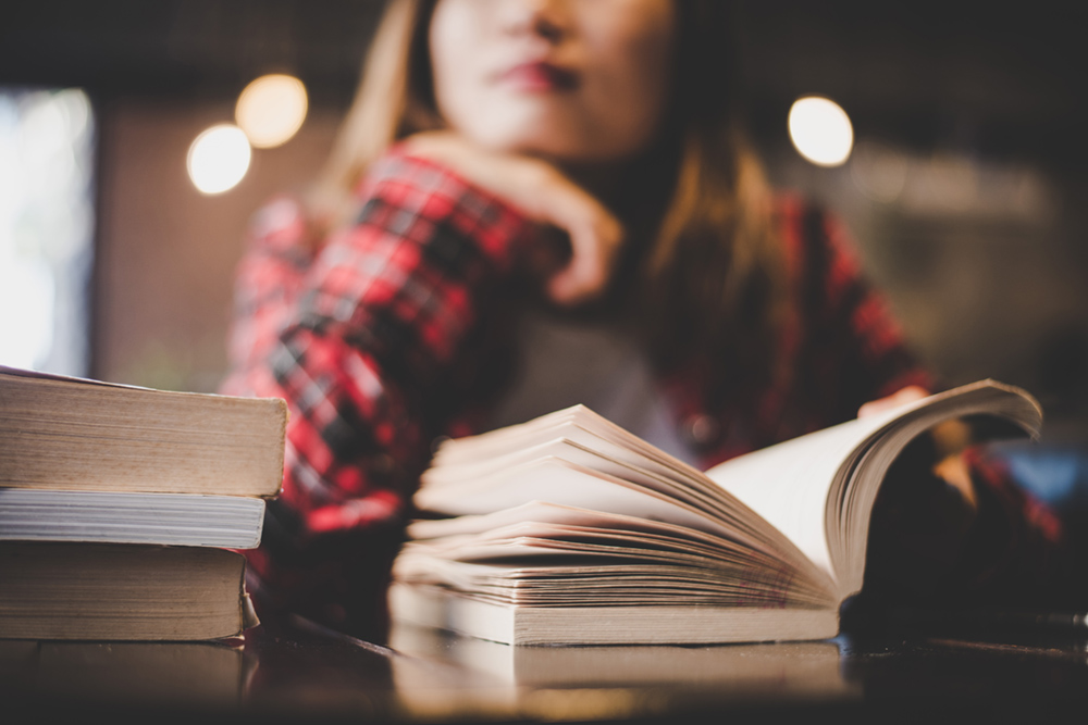 Female in a flannel shirt with chin on hand, looking into the distance, with a book open on the table. Soft focus, maybe a library